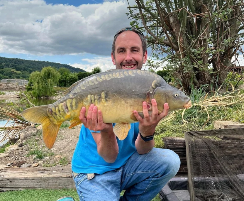 Angler holding a mirror carp beside a landing net and unhooking mat